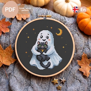 May include: A circular wooden embroidery hoop containing a cross stitch pattern on black fabric. The design features a white ghost with a smiling face holding a black cat with green eyes and a yellow star collar, surrounded by small yellow stars and a crescent moon.