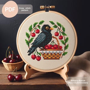 May include: Cross-stitch pattern featuring a blackbird perched above a basket of cherries, surrounded by cherry branches. The design is displayed in a wooden embroidery hoop, with the text "PDF" and "Cross stitch pattern".