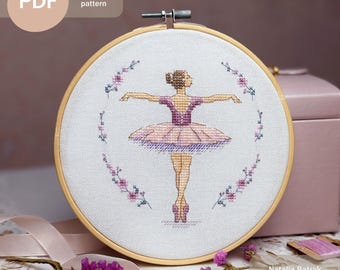 May include: Cross stitch pattern PDF of a ballerina in a pink tutu with a floral wreath around her. The ballerina is in a pose with her arms outstretched. The pattern is in a wooden embroidery hoop.