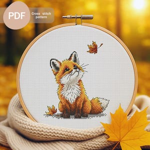Puede incluir: Un patrón de punto de cruz de un zorro en un bastidor de bordado de madera. El zorro es naranja y blanco, con una expresión alegre, y está rodeado de hojas de otoño. El texto "PDF Cross stitch pattern" es visible.