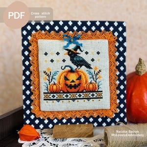 Könnte beinhalten: Kreuzstichmuster mit einem schwarzen Raben, der auf einem Jack-o'-Lantern mit einem gruseligen Gesicht sitzt. Das Design ist von orangefarbenen Kürbissen und Blättern umgeben, und der Hintergrund ist ein weißer Stoff mit einem blau-weißen Polka-Dot-Rand.