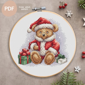 Könnte beinhalten: Ein Kreuzstichmuster mit einem braunen Teddybär, der einen roten Weihnachtsmann-Hut und einen roten Mantel trägt und eine Geschenkbox hält. Der Bär sitzt auf einem violetten Hintergrund mit weißen Schneeflocken. Das Muster ist auf einem weißen Stoff mit einem Holzreifen.