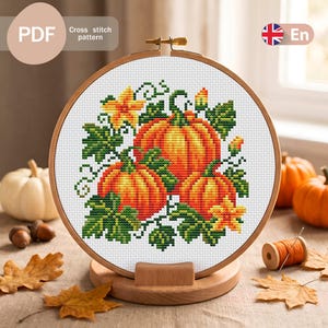 Puede incluir: Un bastidor de bordado circular de madera que contiene un patrón de punto de cruz de tres calabazas naranjas con hojas verdes y flores amarillas. El diseño está impreso en tela blanca. El texto PDF, Cross stitch pattern y En es visible.