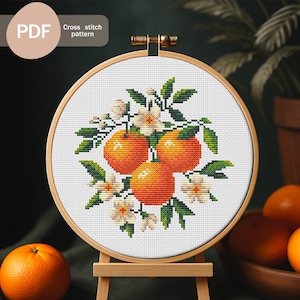 May include: A cross-stitch pattern of oranges and blossoms, displayed in a wooden embroidery hoop. The design features three orange oranges, white flowers, and green leaves. The text "PDF" and "Cross stitch pattern" are visible.