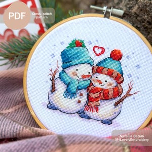May include: Cross stitch pattern of two snowmen in love. The snowmen are wearing blue and red scarves and hats. A red heart is above them. The pattern is on a white background with a wooden hoop.