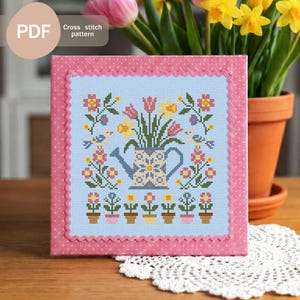 Peut inclure: Un motif de point de croix encadré avec un motif floral, comprenant un arrosoir rempli de fleurs. Le motif est sur un fond bleu clair avec une bordure rose. Le texte "Cross stitch pattern" est visible.