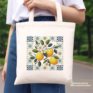 May include: Cream-colored tote bag featuring a cross-stitch design of lemons, white blossoms, and green leaves. The design is set against a backdrop of blue, yellow, and white geometric patterns. The bag is held by a person.