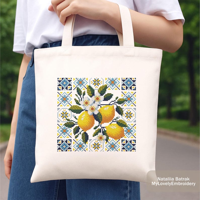May include: Cream-colored tote bag featuring a cross-stitch design of lemons, white blossoms, and green leaves. The design is set against a backdrop of blue, yellow, and white geometric patterns. The bag is held by a person.