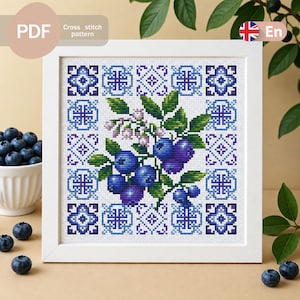 Pode incluir: Um padrão de ponto cruz apresentando um cacho central de mirtilos com folhas verdes e pequenas flores brancas, emoldurado por uma borda geométrica azul e branca, apresentado em uma moldura quadrada branca com etiquetas de texto PDF e En.