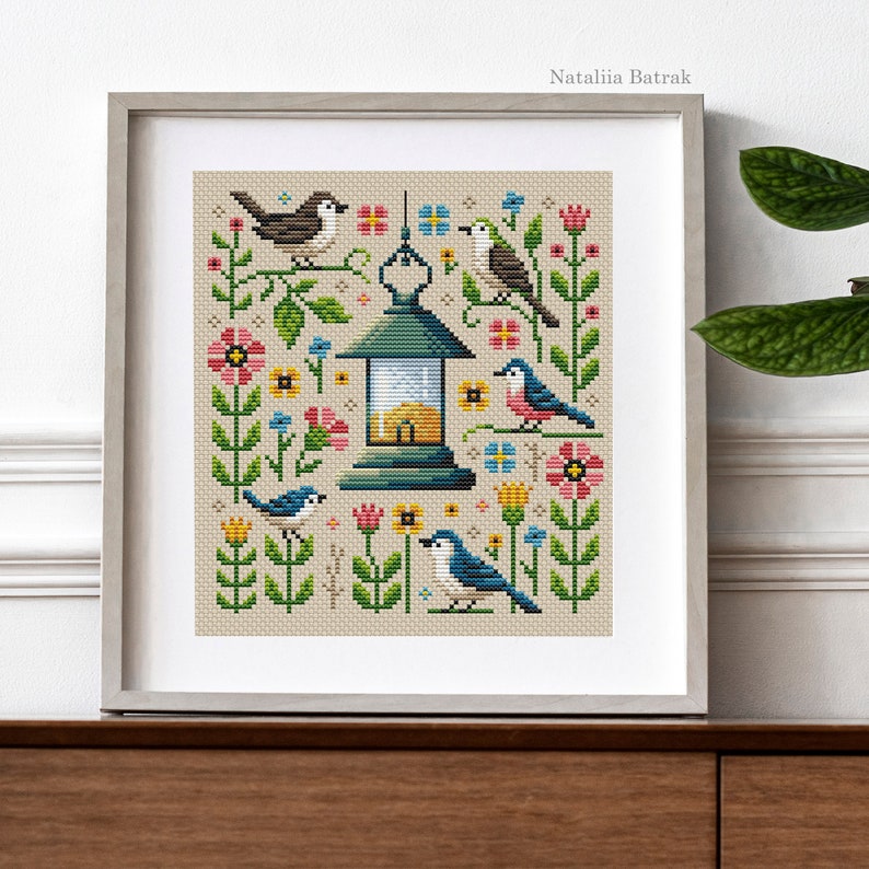 May include: A cross-stitch pattern featuring a lantern with a birdhouse inside, surrounded by colorful flowers and birds. The pattern is framed in a light gray frame.