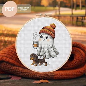 Puede incluir: Un patrón de punto de cruz en un aro de madera presenta un fantasma de dibujos animados con un gorro naranja y sosteniendo una taza de café, paseando a un perro salchicha. El texto "PDF Cross stitch pattern" está en la esquina superior izquierda. El fondo está borroso.