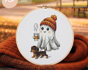 May include: A cross-stitch pattern in a wooden hoop features a cartoon ghost wearing an orange beanie and holding a coffee cup, walking a dachshund. The text "PDF Cross stitch pattern" is in the upper left corner. The background is blurred.
