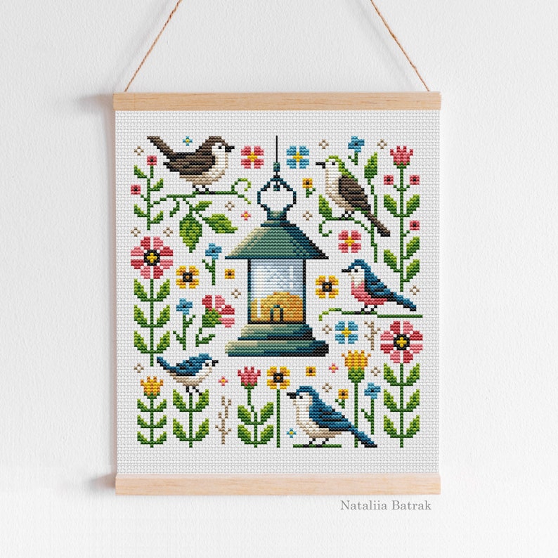 May include: Cross stitch pattern featuring a lantern with a yellow window, surrounded by colorful flowers and birds. The pattern includes blue, green, yellow, red, and pink colors. The text "Natalia Batrak" is at the bottom of the pattern.