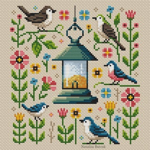 May include: A cross-stitch pattern featuring a lantern with a glowing light inside, surrounded by colorful flowers and birds. The birds are blue, brown, and pink, and the flowers are yellow, pink, and blue. The background is a light beige.