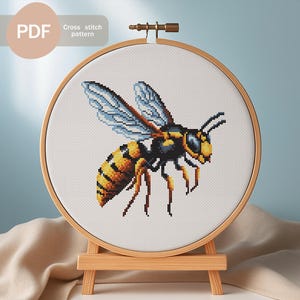 Könnte beinhalten: Ein Kreuzstichmuster einer Wespe, präsentiert in einem hölzernen Stickrahmen auf einer kleinen Staffelei. Die Wespe ist mit gelbem, schwarzem und blauem Garn gestickt. Der Text "PDF" und "Cross stitch pattern" befindet sich oben links.
