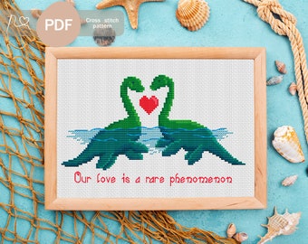 May include: Cross-stitch pattern showing two green dinosaur-like creatures with heart-shaped necks. The design includes blue water lines and the text "Our love is a rare phenomenon" in red. The pattern is framed in a light wood. The words "PDF" and "Cross stitch pattern" are also visible.