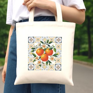 Puede incluir: Bolso tote de color crema con un dise&ntilde;o de punto de cruz que presenta tres naranjas, flores blancas y hojas verdes. El dise&ntilde;o est&aacute; enmarcado por un patr&oacute;n cuadrado en azul, amarillo y blanco. El bolso tiene dos asas color crema.