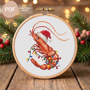 Pode incluir: Um padrão de ponto cruz de um camarão usando um gorro de Papai Noel e luzes de Natal, em um bastidor de bordado de madeira. O texto "Cross stitch pattern" é visível. O design está em um fundo branco.
