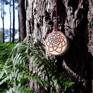 May include: A wooden pendant with a Celtic knot design carved into it. The pendant is hanging from a red string on a tree trunk.