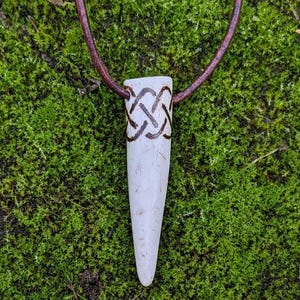 May include: A white bone pendant with a brown Celtic knot design, hanging from a brown leather cord.