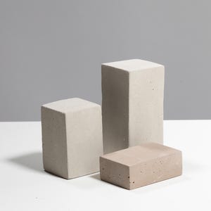 May include: Three rectangular blocks made of light gray and beige concrete with a porous, textured surface. Two blocks are positioned vertically at different heights, while the third block lies horizontally in the foreground.