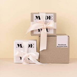 Puede incluir: Tres cajas de regalo, dos blancas y una marr&oacute;n, est&aacute;n apiladas. Las cajas blancas tienen el texto "MADE YOU" en negro. La caja marr&oacute;n tiene "Handmade BETONVTON" en negro. Cada caja est&aacute; atada con una cinta de raso rosa claro.