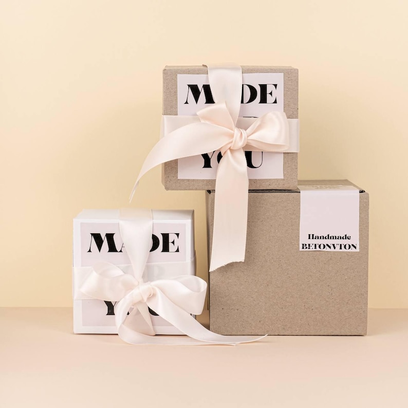 Puede incluir: Tres cajas de regalo, dos blancas y una marr&oacute;n, est&aacute;n apiladas. Las cajas blancas tienen el texto "MADE YOU" en negro. La caja marr&oacute;n tiene "Handmade BETONVTON" en negro. Cada caja est&aacute; atada con una cinta de raso rosa claro.
