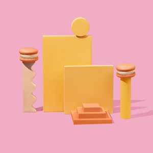Könnte beinhalten: Ein Stillleben mit orangefarbenen Macarons auf gelben und beigen geometrischen Formen vor rosa Hintergrund. Die Formen umfassen einen Zylinder, eine wellenförmige Säule und gestapelte Quadrate. Die Macarons haben eine cremige Füllung.