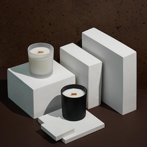 Pode incluir: Duas velas com pavios de madeira s&atilde;o exibidas em blocos geom&eacute;tricos brancos contra um fundo marrom. Uma vela est&aacute; em um recipiente de vidro fosco, a outra em um recipiente preto. As velas s&atilde;o preenchidas com cera branca cremosa.