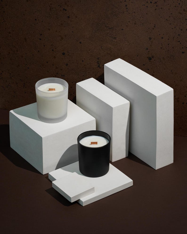 Pode incluir: Duas velas com pavios de madeira s&atilde;o exibidas em blocos geom&eacute;tricos brancos contra um fundo marrom. Uma vela est&aacute; em um recipiente de vidro fosco, a outra em um recipiente preto. As velas s&atilde;o preenchidas com cera branca cremosa.