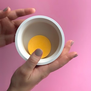 Puede incluir: Un cuenco redondo de cer&aacute;mica blanca con un c&iacute;rculo amarillo en su interior, sostenido por manos sobre un fondo rosa. El interior del cuenco es de color beige claro, creando contraste con el c&iacute;rculo amarillo brillante.