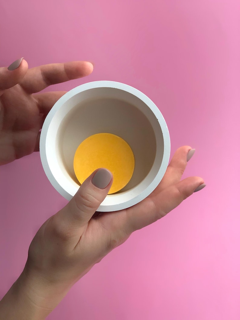 Puede incluir: Un cuenco redondo de cer&aacute;mica blanca con un c&iacute;rculo amarillo en su interior, sostenido por manos sobre un fondo rosa. El interior del cuenco es de color beige claro, creando contraste con el c&iacute;rculo amarillo brillante.