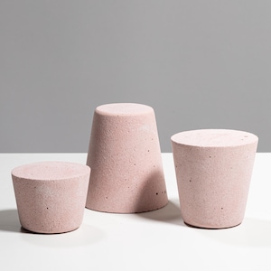May include: Three light pink concrete objects with a porous, textured surface. The set includes one short, wide cylindrical piece, one taller, narrower tapered piece, and one medium-height cylindrical piece, all featuring a flat top surface.