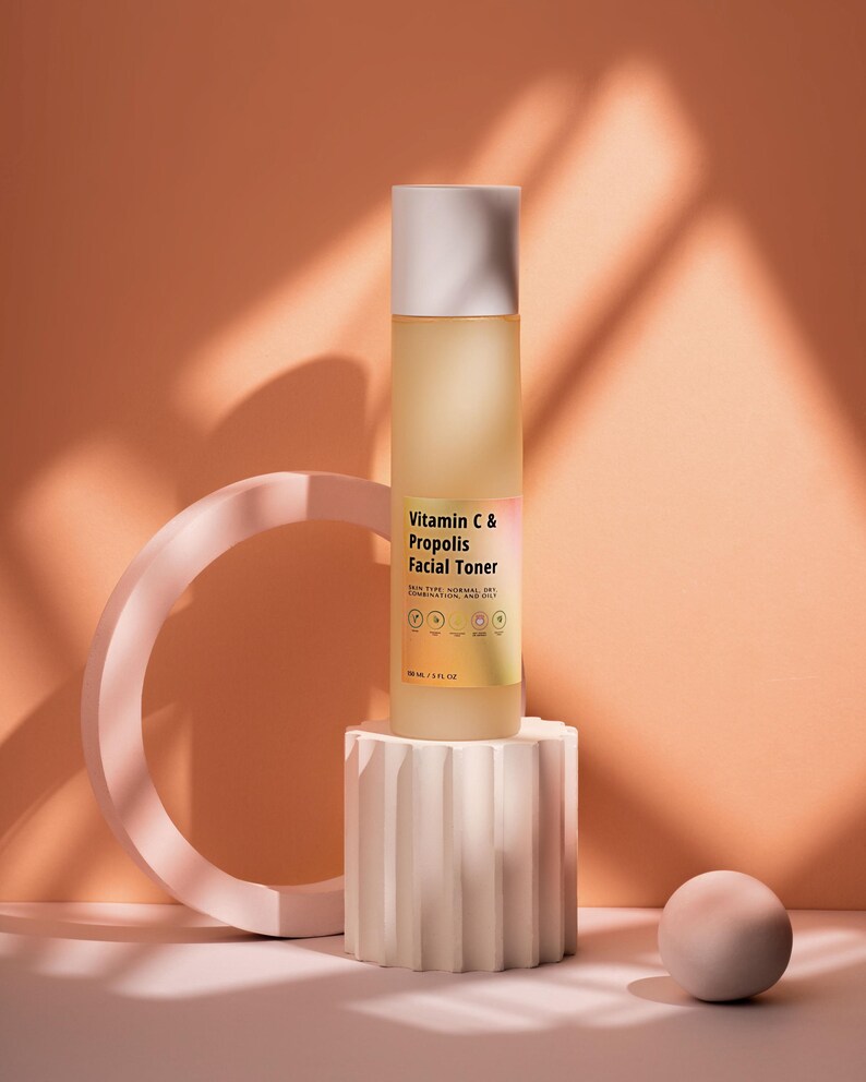 Puede incluir: Una botella de vidrio transparente de t&oacute;nico facial de vitamina C y prop&oacute;leo con tapa blanca, colocada sobre un pedestal blanco texturizado. La botella est&aacute; etiquetada con informaci&oacute;n del producto. El fondo es de color melocot&oacute;n c&aacute;lido con un dise&ntilde;o geom&eacute;trico.