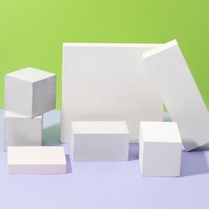 Könnte beinhalten: Anordnung weißer geometrischer Formen, darunter Würfel und rechteckige Prismen, auf einer hellvioletten Oberfläche vor einem grünen Hintergrund. Die Formen sind minimalistisch und schaffen eine moderne Ästhetik.