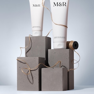 May include: Two white shampoo tubes with the text "M&R" and "SHAMPOO FOR CURLY HAIR WITH BAOBAB OIL AND PHOSPHOLIPIDS" are displayed on gray concrete blocks. The tubes are adorned with twine, and a spool of twine is also present.