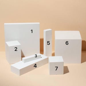 Puede incluir: Una colección de formas geométricas de espuma blanca, que incluyen prismas rectangulares, cubos y un trapecio, dispuestas sobre un fondo neutro. Las formas varían en tamaño y están diseñadas para fotografía de productos o exhibición.