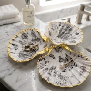 Könnte beinhalten: Drei dekorative Muschelschalen mit einem schwarz-weißen Blumen- und Schmetterlingsdesign. Die Ränder sind mit Gold verziert. Ringe und eine Halskette mit einer Perle werden auf den Schalen präsentiert. Die Schalen stehen auf einer Marmorarbeitsplatte.