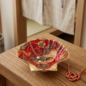 Könnte beinhalten: Eine dekorative Muschelschale mit leuchtend rotem Inneren, verziert mit einem Vogel- und Blumenmuster und einem goldfarbenen Rand. In der Schale liegen eine goldene Halskette mit Perlenanhänger, ein goldener Ring und eine kleine goldene Anstecknadel.