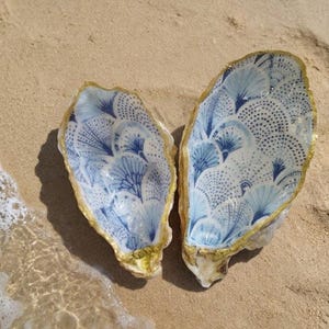 Peut inclure: Deux coquilles d'huîtres décoratives avec un motif floral bleu et blanc et une bordure dorée, posées sur une plage de sable. Les coquilles sont ouvertes, révélant le motif détaillé. La scène est près du bord de l'eau.