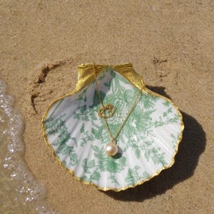 Peut inclure: Un collier en or avec un pendentif perle est posé dans une coquille Saint-Jacques décorative. Le plat a un motif floral blanc et vert avec une bordure dorée. La coquille est sur du sable près de l'eau.