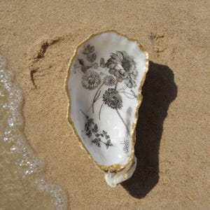Peut inclure: Coquille d'huître blanche avec un bord doré, ornée d'un motif floral noir et blanc. Posée sur une plage de sable, avec l'eau en arrière-plan.