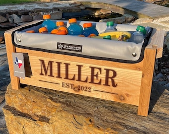 Op de afbeelding: Een houten koelbox met een grijze voering, gepersonaliseerd met de naam "MILLER" en de datum "EST. 2022". De koelbox is gevuld met drankjes en een banaan. De koelbox staat op een grote rots.