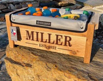 May include: A wooden ice chest with a grey liner, personalised with the name "MILLER" and the text "EST. 2022". The chest is filled with drinks and fruit, and is sitting on a large rock.