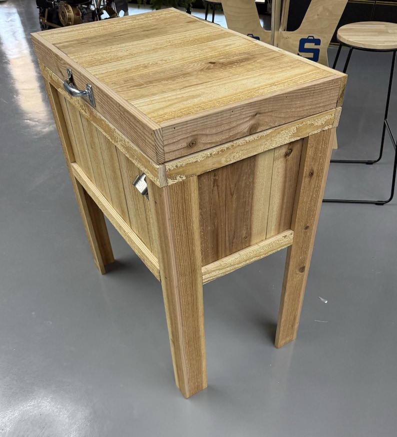 以下が含まれることがあります： 金属製の取っ手と蝶番付きの蓋が付いた木製クーラー。クーラーは自然な木でできており、薄い茶色の仕上げになっています。素朴な外観で、屋外でのエンターテイメントに最適です。