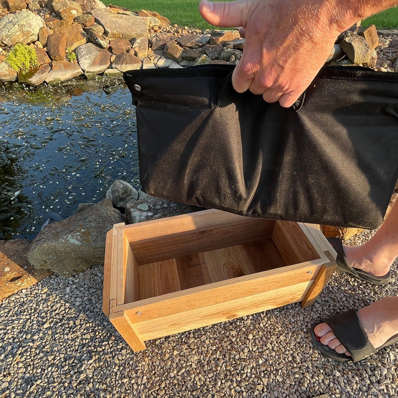 May include: A wooden planter box with a black fabric liner. The liner is being lifted out of the box by a hand.