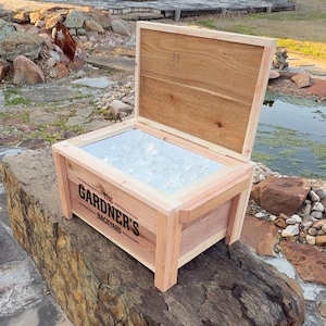 May include: A wooden cooler with a hinged lid, open to reveal a layer of ice. The cooler is labeled "The Gardner's Backyard".