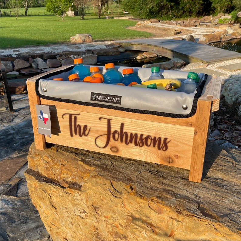 Cedar Wood ice chest with Removable insulated liner inside, Sitting on a rock with a personalization engraved across the front of the cooler