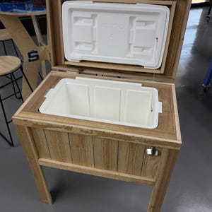 以下が含まれることがあります： 白いイグルークーラーが入った木製クーラースタンド。クーラーは開いており、蓋は持ち上げられています。スタンドは木製の上部と脚、側面に金属製のボトルオープナーが付いています。