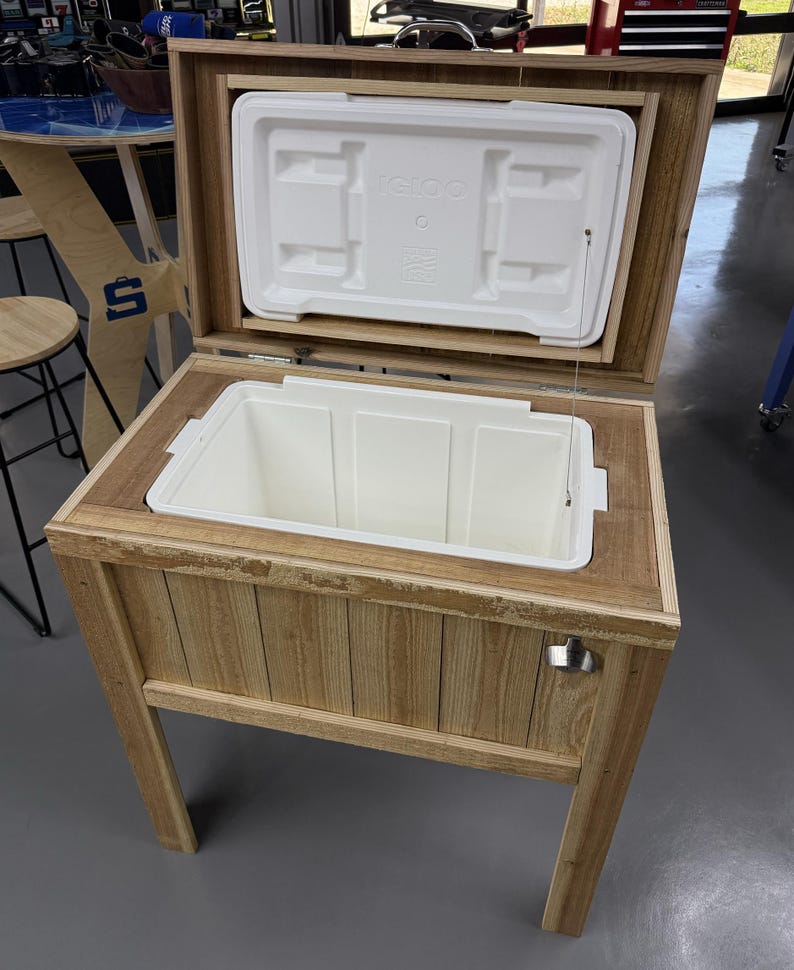 以下が含まれることがあります： 白いイグルークーラーが入った木製クーラースタンド。クーラーは開いており、蓋は持ち上げられています。スタンドは木製の上部と脚、側面に金属製のボトルオープナーが付いています。